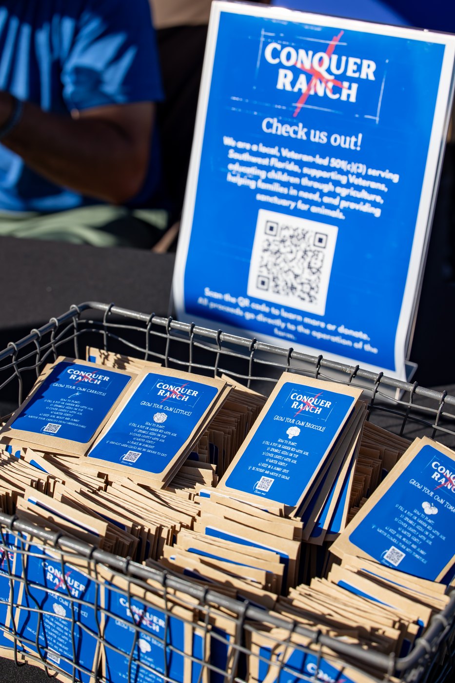 Seed packets with blue Conquer Ranch labels are arranged in a basket on the event table.
