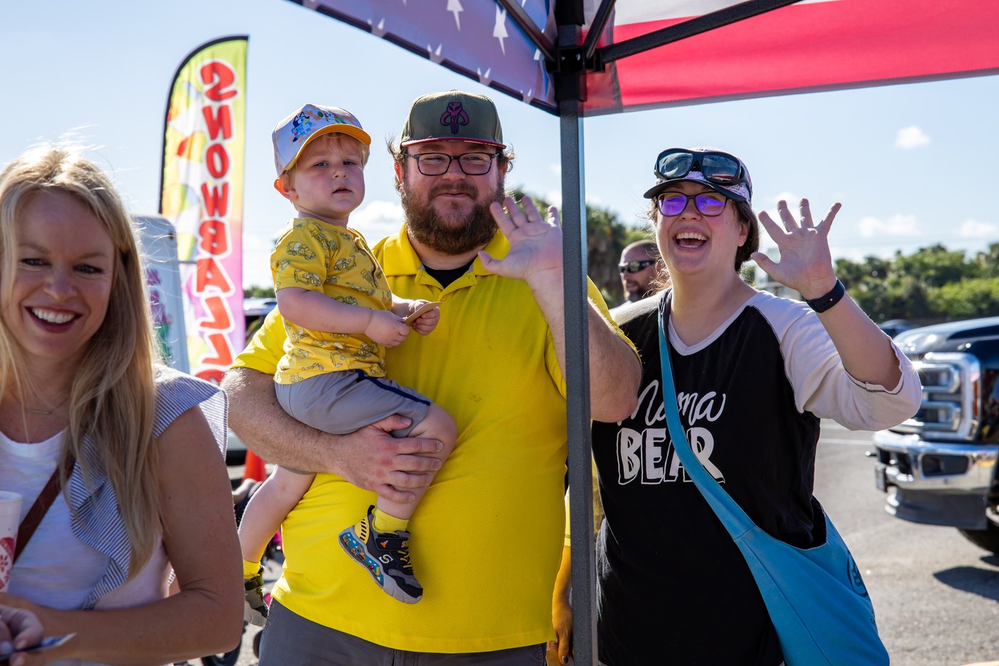 A smiling adult holds a young child while another visitor waves beneath the event canopy.