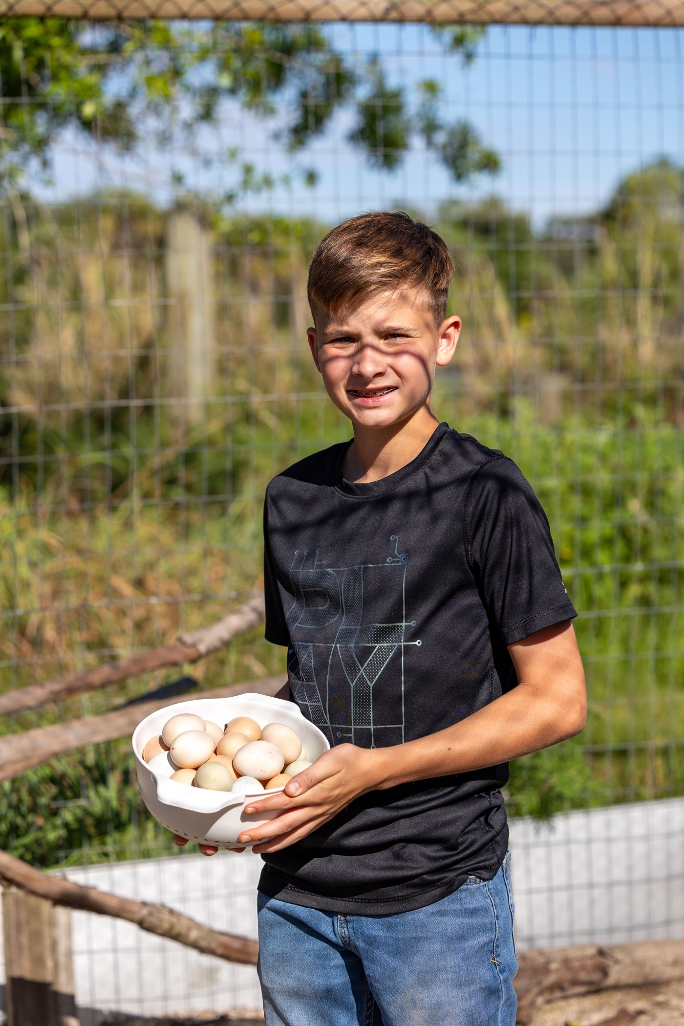 A child holding a bowl of eggs beside a ranch enclosure