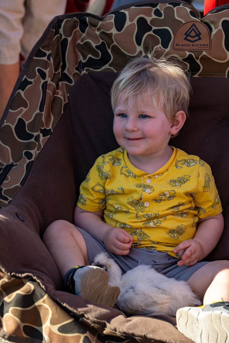 A smiling toddler sits in a camp chair with a fluffy white chicken resting near their legs