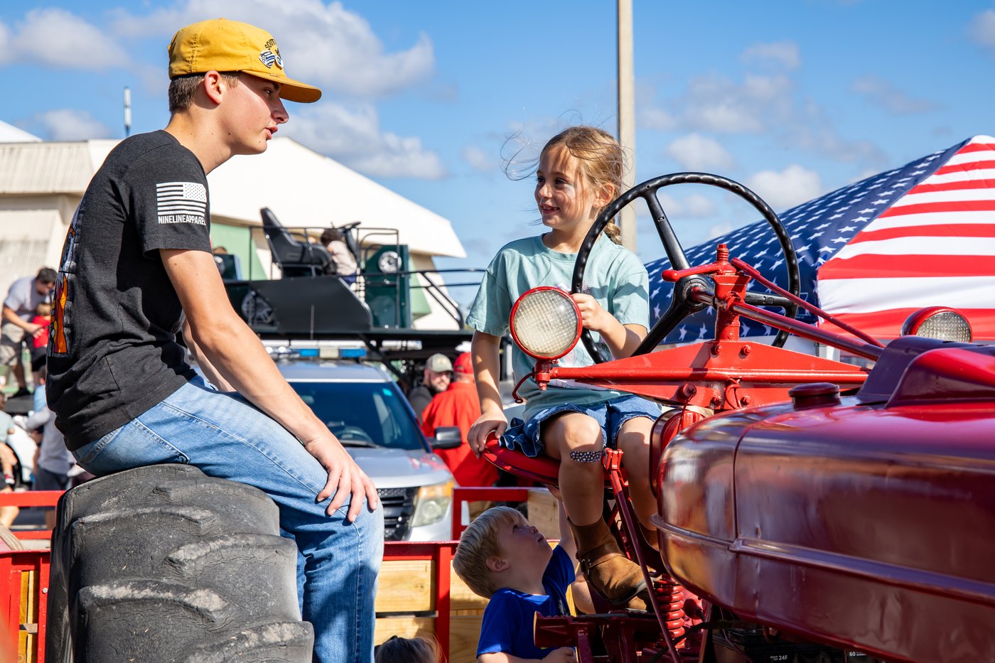 A child seated on a red tractor smiles at a person sitting beside its rear wheel